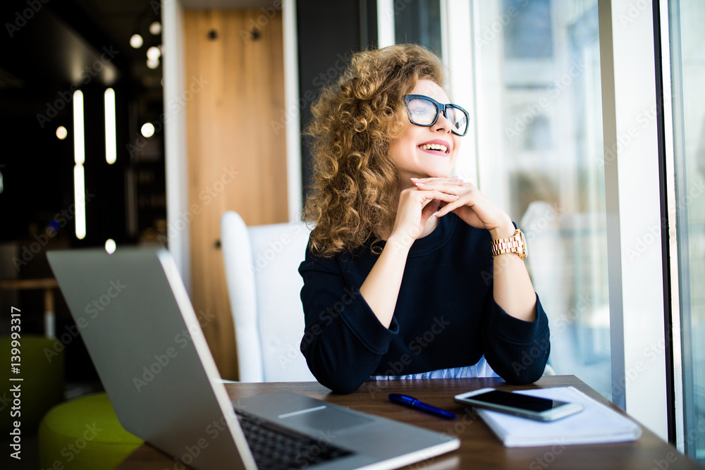 Thoughtful young beautiful woman holding hand on chin and looking through a window while sitting at her working place Thoughtful young beautiful woman holding hand on chin and looking through a window while sitting at her working place