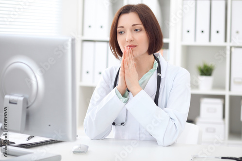 Young brunette female doctor sitting at a desk and working on the computer at the hospital office. Health care, insurance and help concept. Physician ready to examine patient
