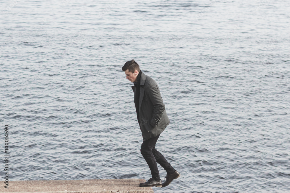 handsome young man in coat walking near water