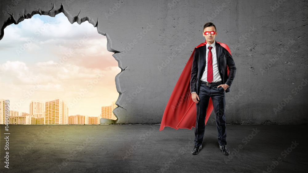 A businessman in a hero red cape with a hand in his pocket on concrete background with a hole to the city view.