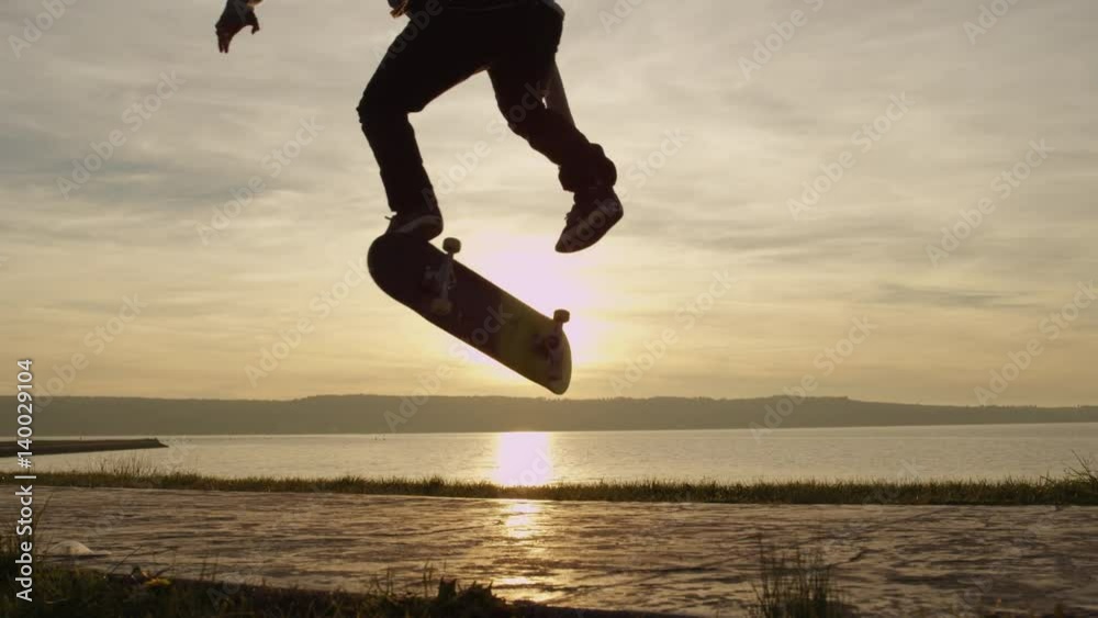SLOW MOTION CLOSE UP, DOF: Silhouetted skateboarder skateboarding and jumping kickflip trick on walking path along the ocean coastline against golden setting sun. Skater riding skateboard doing tricks