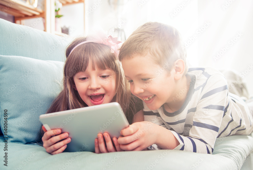Happy siblings lying on sofa at home and playing with tablet together Happy siblings lying on sofa at home and playing with tablet together