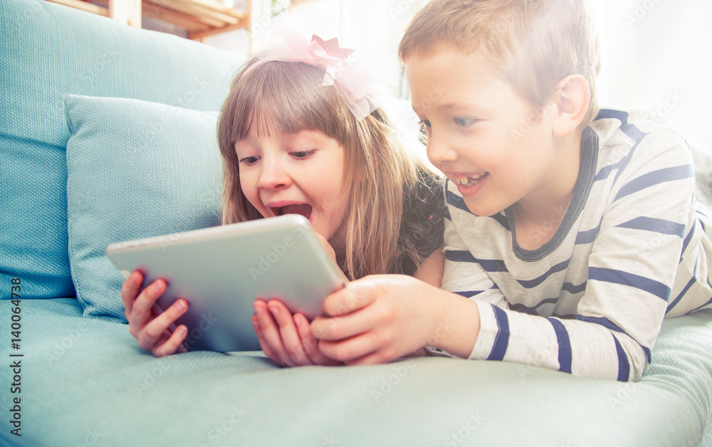 Happy siblings lying on sofa at home and playing with tablet together Happy siblings lying on sofa at home and playing with tablet together