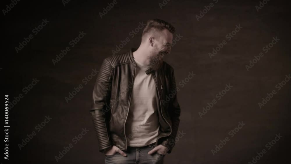 Handsome man with a beard in a black leather jacket posing in the studio on a black background.A man with a beard laughs Handsome man with a beard in a black leather jacket posing in the studio on a black background.A man with a beard laughs