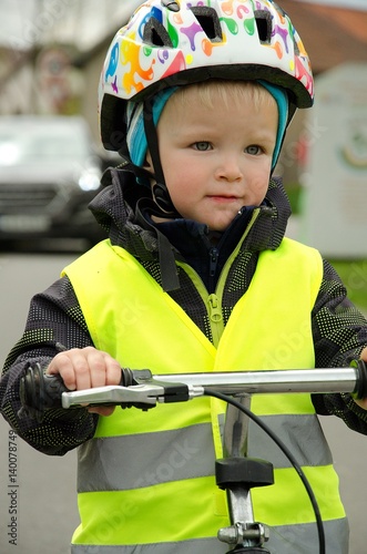 Little child learns to ride a bike on the road. There is a car in the background. Boy is marked by yellow reflective vest and helmet because of visibility and safety.