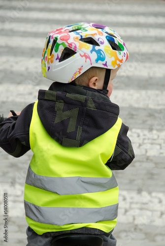 Little child rides a bike across the zebra crossing.  Boy is marked by yellow reflective vest and helmet because of visibility and safety.