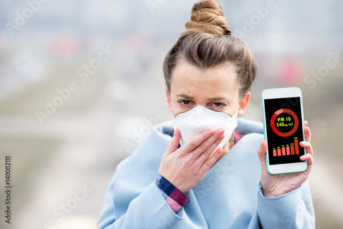 Woman in protective mask holding smart phone with air polution measurement of PM10 outdoors photo