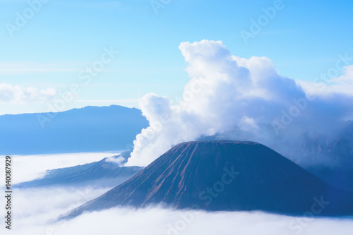 White smoke coming out of volcanoes surrounded by white clouds of mist and a clear blue sky seen at a distance in the afternoon at National Park in East Java, Indonesia.