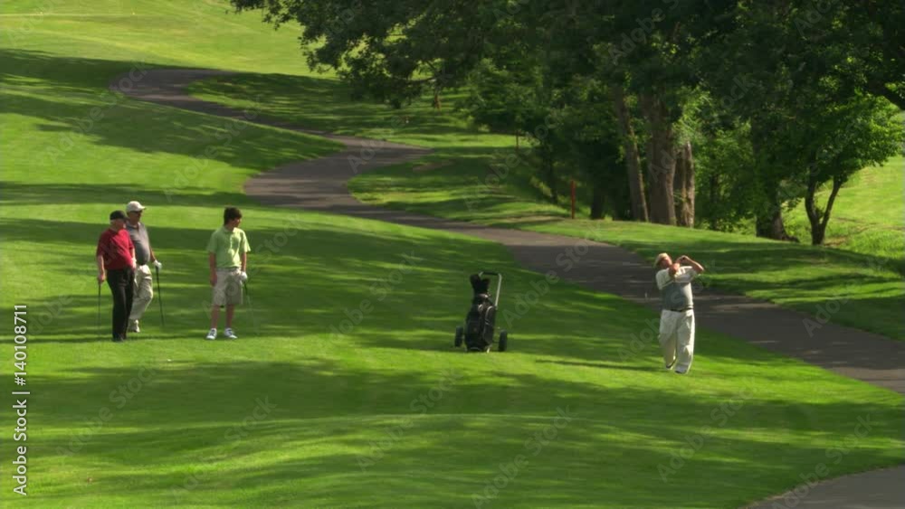 Three golfers standing in shade and watching a third hitting a ball near cart path Three golfers standing in shade and watching a third hitting a ball near cart path