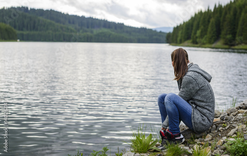 Beautiful young woman relaxing by the mountain lake sitting on the edge of lakeshore looking aside. Enjoing nature