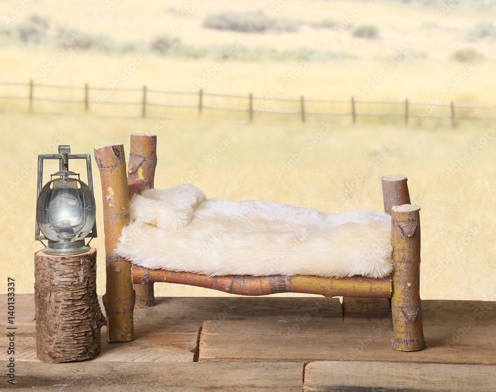 A newborn backdrop background photography prop with a rustic log bed, a railroad lantern on a nightstand, a faux cream bedspread and a yellow country meadow with a fence in the background.