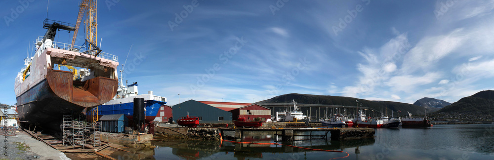 Panorama einer Werft im Hafen von Tromso, Norwegen, mit Schiff auf dem Trockendock, Ölsperre, Lagerhallen, Trawler, Fischkutter und der Tromsøbrua im Hintergrund Panorama einer Werft im Hafen von Tromso, Norwegen, mit Schiff auf dem Trockendock, Ölsperre, Lagerhallen, Trawler, Fischkutter und der Tromsøbrua im Hintergrund