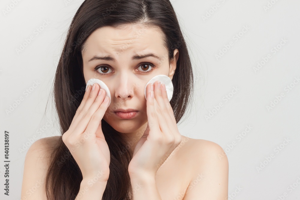 The brunette on a white background with cream on her face, clean face, sponges in hand sad The brunette on a white background with cream on her face, clean face, sponges in hand sad