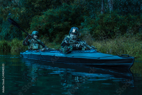 Special forces men with painted faces in camouflage uniforms paddling army kayak. Boat moving calmly across the river, diversionary mission, twilight photo