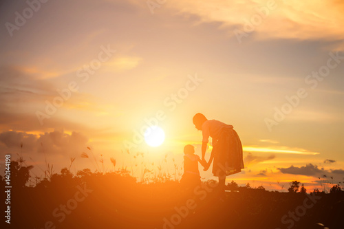 a silhouette of a happy young girl child the arms of his loving mother for a hug, in front of the sunset in the sky on a summer day.