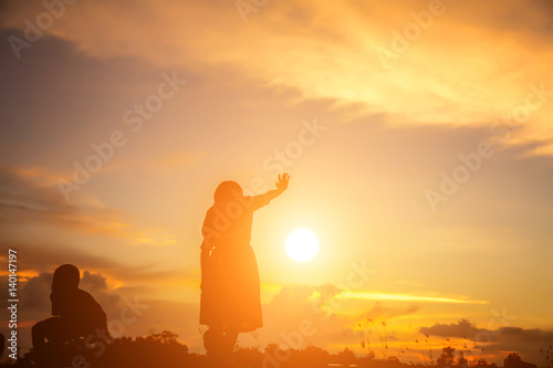 a silhouette of a happy young girl child the arms of his loving mother for a hug  in front of the sunset in the sky on a summer day.