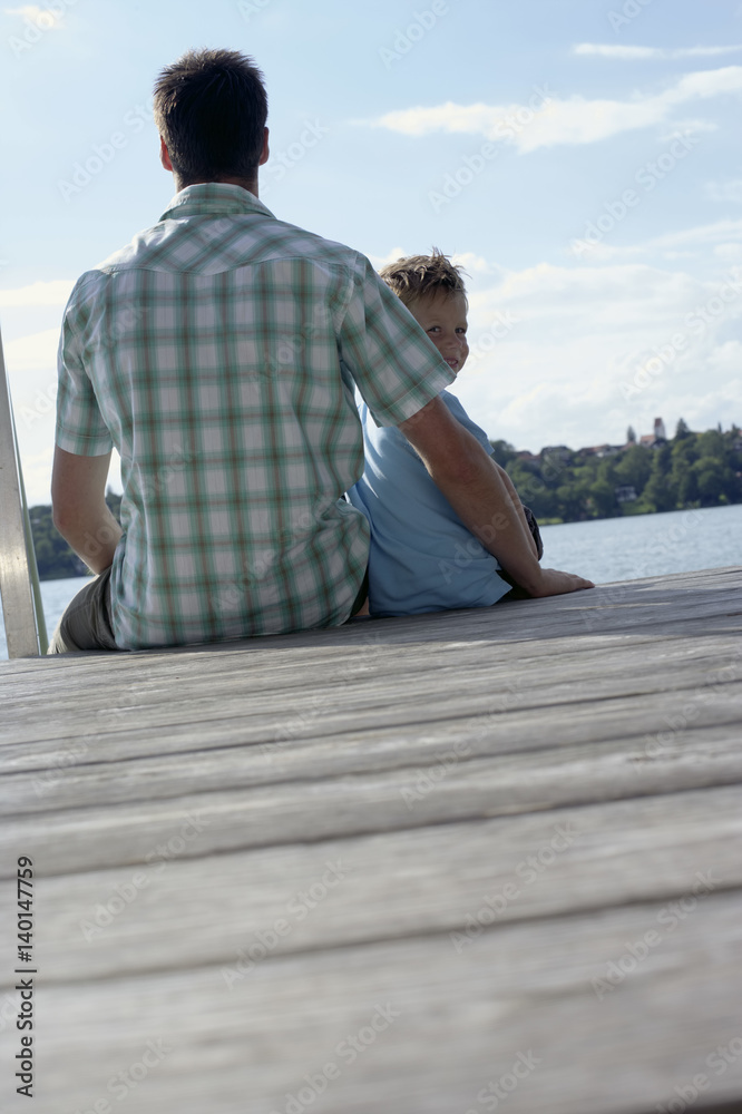 Fototapeta premium Father and son sitting on a wooden footbridge next to each other, selective focus