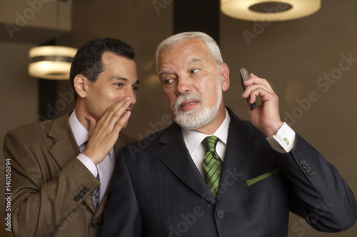 Businessman whispering in mature businessman's ear holding a mobile phone