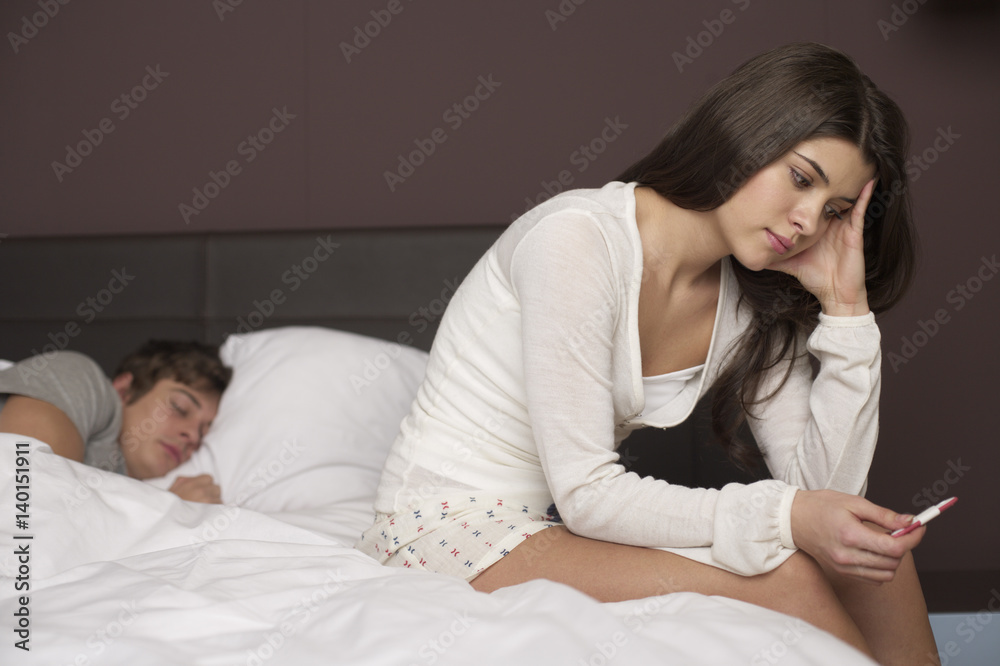 Young brunette woman is sitting at the edge of a bed staring at a pregnancy test Young brunette woman is sitting at the edge of a bed staring at a pregnancy test