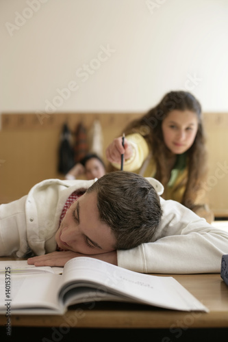 Teenage boy sleeping in a classroom, teenage girl tapping him