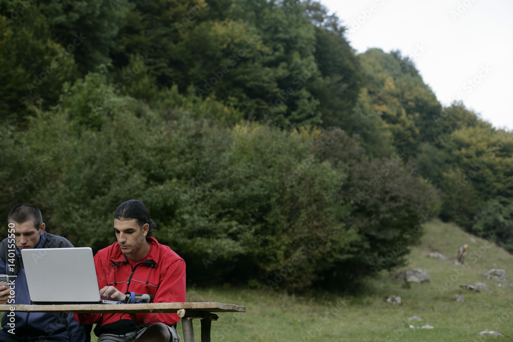 Young man with black hair with a laptop is sitting next to a man with a cell phone his hand, both on a bench outside Young man with black hair with a laptop is sitting next to a man with a cell phone his hand, both on a bench outside