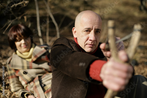 Mature man using a slingshot, woman lying in background