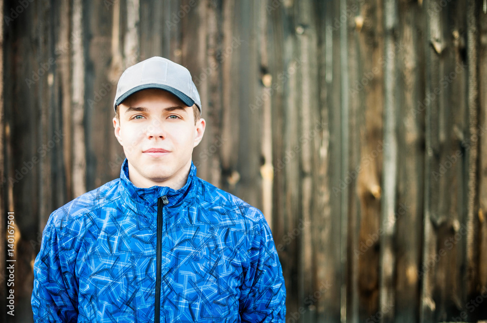 Young attractive guy on the background of a wooden fence, wearing a cap and a blue sports jacket