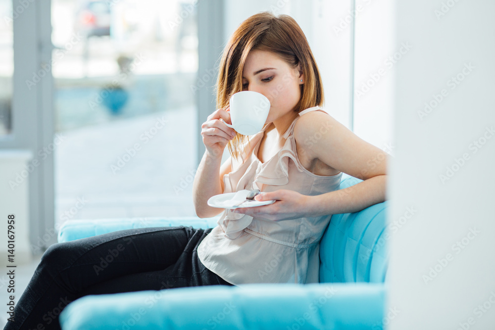 Portrait of female visitor waiting for a doctor in hall of clinic while sitting in turquoise sofa and drinking coffee or tea from white cup on bright lighting background. Portrait of female visitor waiting for a doctor in hall of clinic while sitting in turquoise sofa and drinking coffee or tea from white cup on bright lighting background.