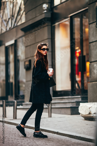 young stylish girl, passing by the Windows.Wearing fashionable glasses and a black coat.Keeps coffee.Abstract looking to the side.