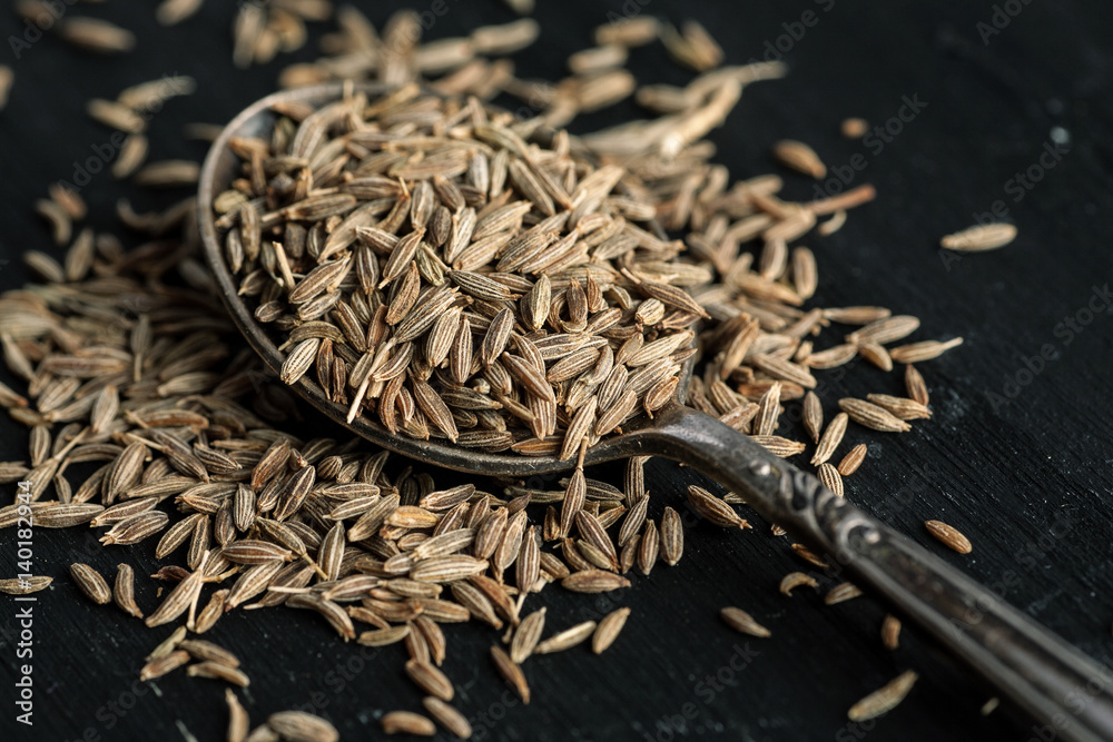 Cumin dry seeds in metal spoon on a black wooden table.