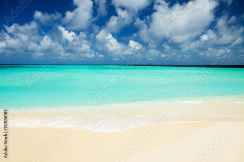 Tropical beach. Ocean waves and cloudy sky background. White sand and crystal-blue sea. Ocean water nature, beach relax. Summer sea vacation. Maldives islands sea background