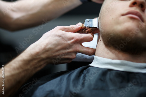 Beard grooming. Side view of young bearded man getting beard haircut by hairdresser while sitting in chair at barbershop