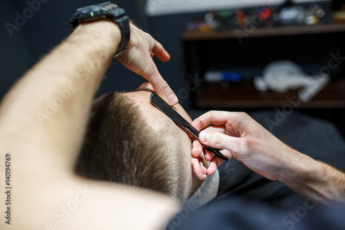 Beard grooming. Side view of young bearded man getting beard haircut by hairdresser while sitting in chair at barbershop