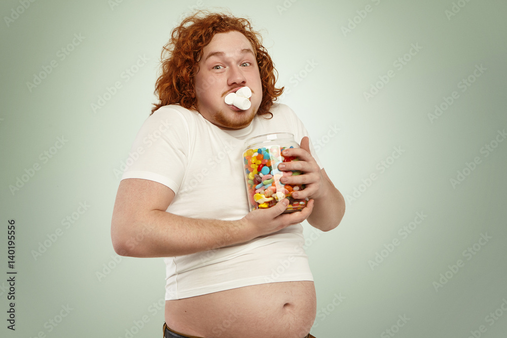 Obesity, gluttony, people and unhealthy lifestyle. Funny fat obese redhead  man in white t-shirt, his belly showing up, holding jar of goodies,  obsessed with food, cramming up mouth with marshmallow Stock Photo |