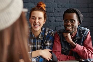 Group of three happy students having fun indoors: beautiful girl with orange hair bun sitting at cafe table close to her African boyfriend, both laughing cheerful and talking to unrecognizable female