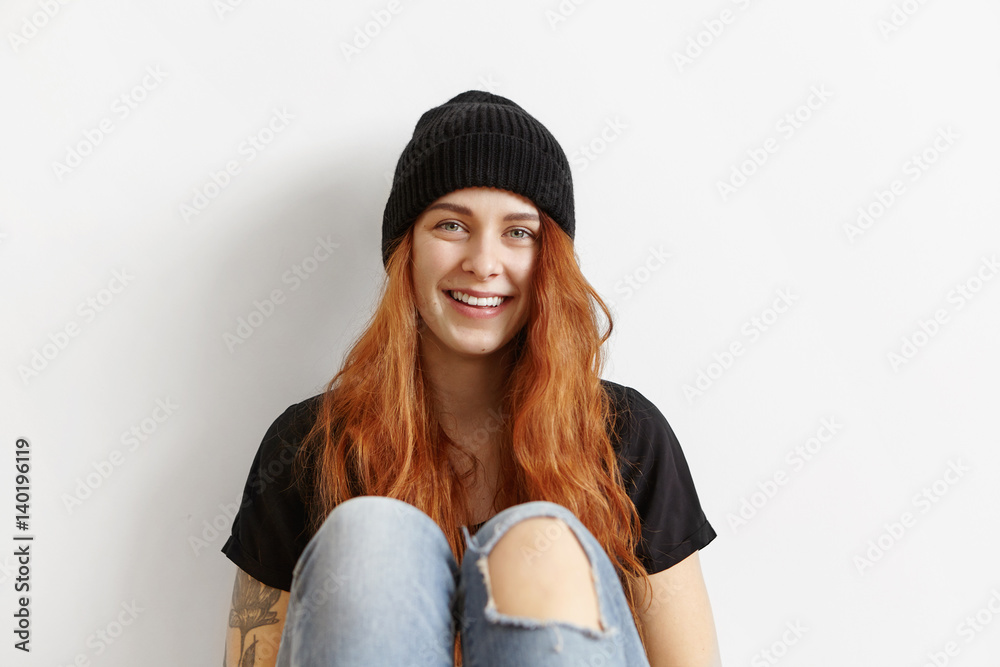 Fashionable female teenager wearing her messy ginger hair loose relaxing indoors, sitting at white wall, dressed in black t-shirt, hat and ragged jeans, having happy look, enjoying free time at home Fashionable female teenager wearing her messy ginger hair loose relaxing indoors, sitting at white wall, dressed in black t-shirt, hat and ragged jeans, having happy look, enjoying free time at home