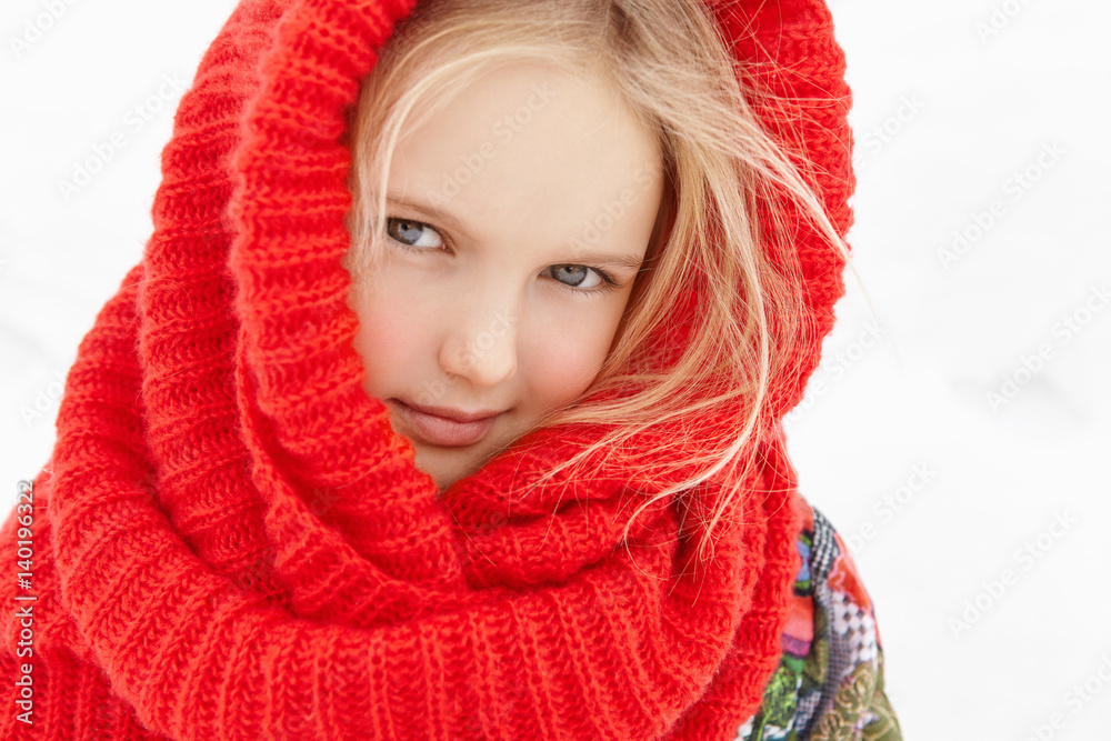 Outdoor view of cute European female child with blonde hair posing outside during short walk in city park in cold winter morning, looking at camera with faint smile, wrapped in red knitted scarf Outdoor view of cute European female child with blonde hair posing outside during short walk in city park in cold winter morning, looking at camera with faint smile, wrapped in red knitted scarf