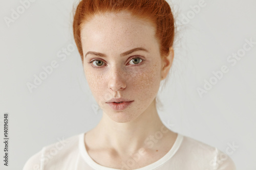 Close up studio shot of beautiful charming redhead European model with healthy freckled skin looking at camera with faint smile, posing indoors against blank wall background, wearing white t-shirt photo