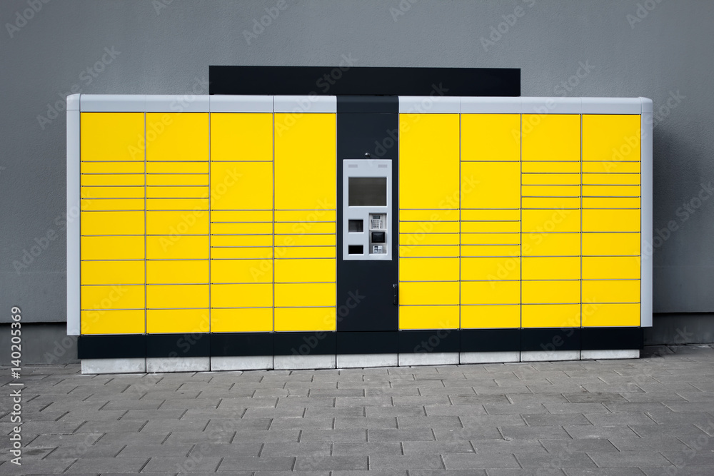 Yellow self-service parcel terminal on the background of gray wall and pavement.