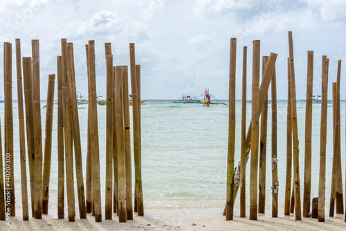 Looking through a bamboo pole barrier fence in a tropical island white sand beach out to a calm crystal clear sea with pump boats anchored in the distance with background focus.
