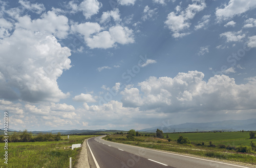 Sunny summer landscape with fluffy clouds in the blue sky and road crossing vast green plains.
