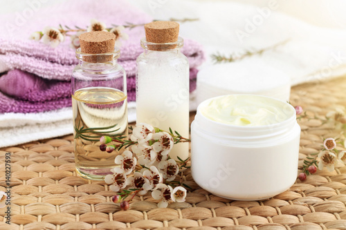 Aroma botanical spa treatment. Bottle of essential oil with flowers, jar of facial moisturizer, towels, sunlight. 