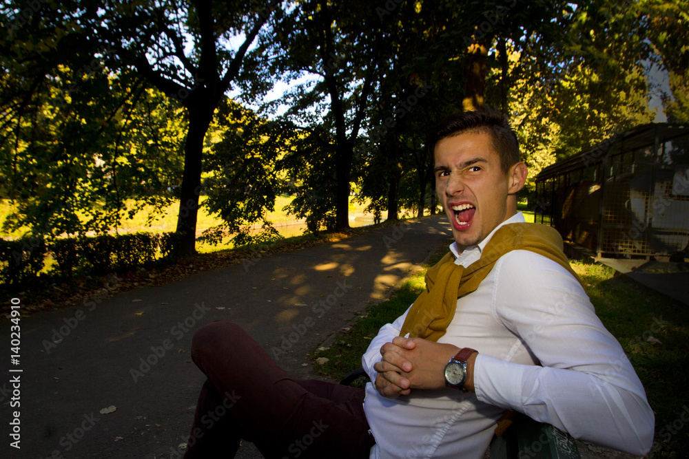 Fototapeta premium Handsome young man dressed in business clothes posing and sitting on a bench,making faces and standing. Young man posing on a sunny day in a park.