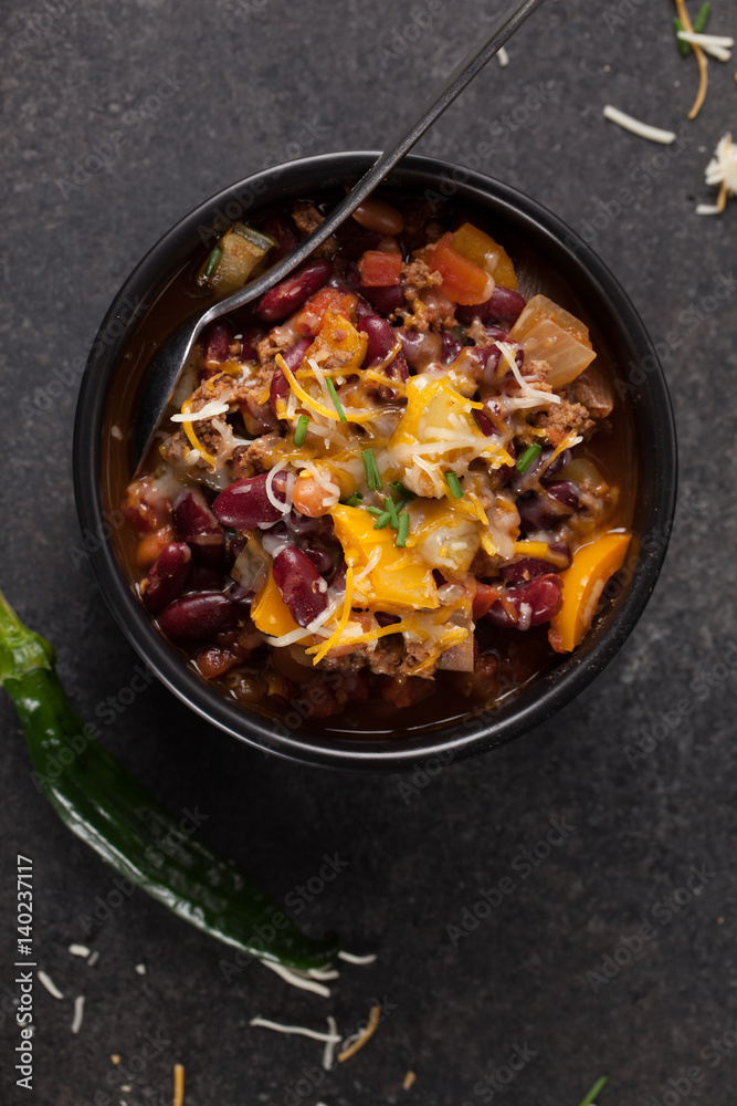 Kidney Bean Chili with tomato chunks and onion topped with cheese in large black bowl above shot