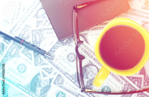 brown glasses and yellow cup of coffee with small notebook and pen on US dollar banknote pattern background © NinelittlePhoto