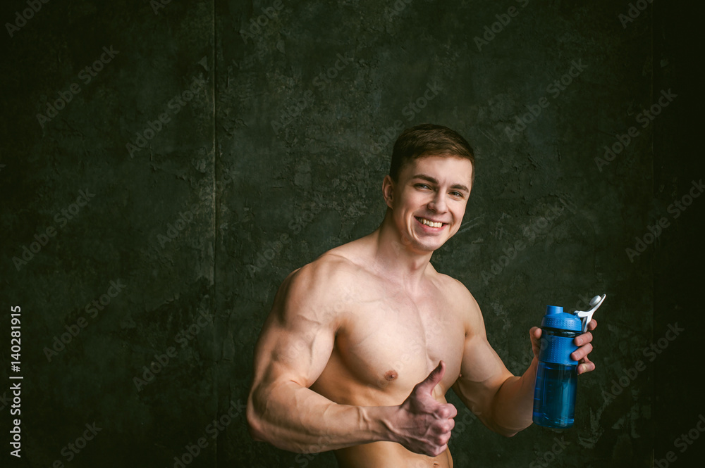 Young sexy men bodybuilder athlete,portrait loft on background of stylized wall,guy model With bare torso Holds in his hand blue sports bottle for drinking during training, show thumbs up and smiling Young sexy men bodybuilder athlete,portrait loft on background of stylized wall,guy model With bare torso Holds in his hand blue sports bottle for drinking during training, show thumbs up and smiling
