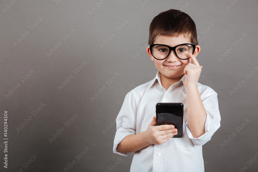 Happy little boy in glasses standing and using mobile phone