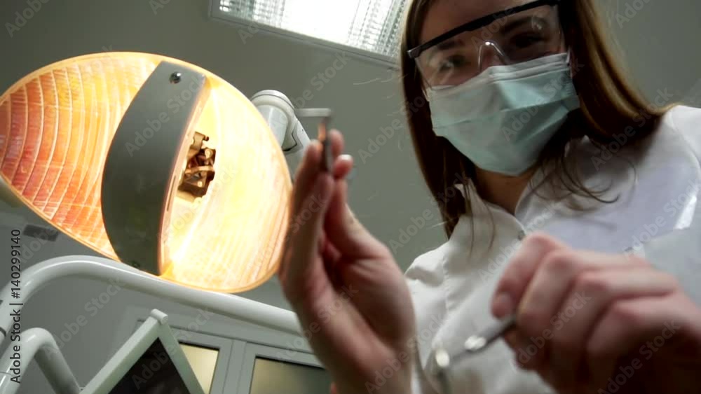 Young Female Dentist in Mask Approaches With Tools, Holding a Mouth Mirror and Dentist's Hook, Dental Lamp Lights Into a Patient's Mouth, Standing Upon a Patient, Looking at Camera, Dentist's Face