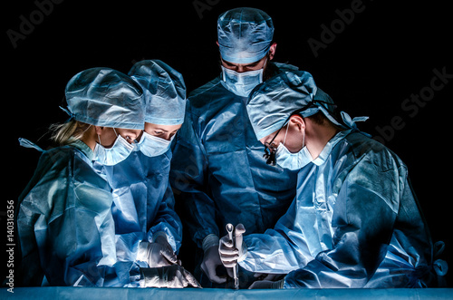 The doctor makes sternotomy during a thoracic operation. Doctors are dressed in blue surgical suits, on their faces they have medical masks, and on their heads surgical caps.