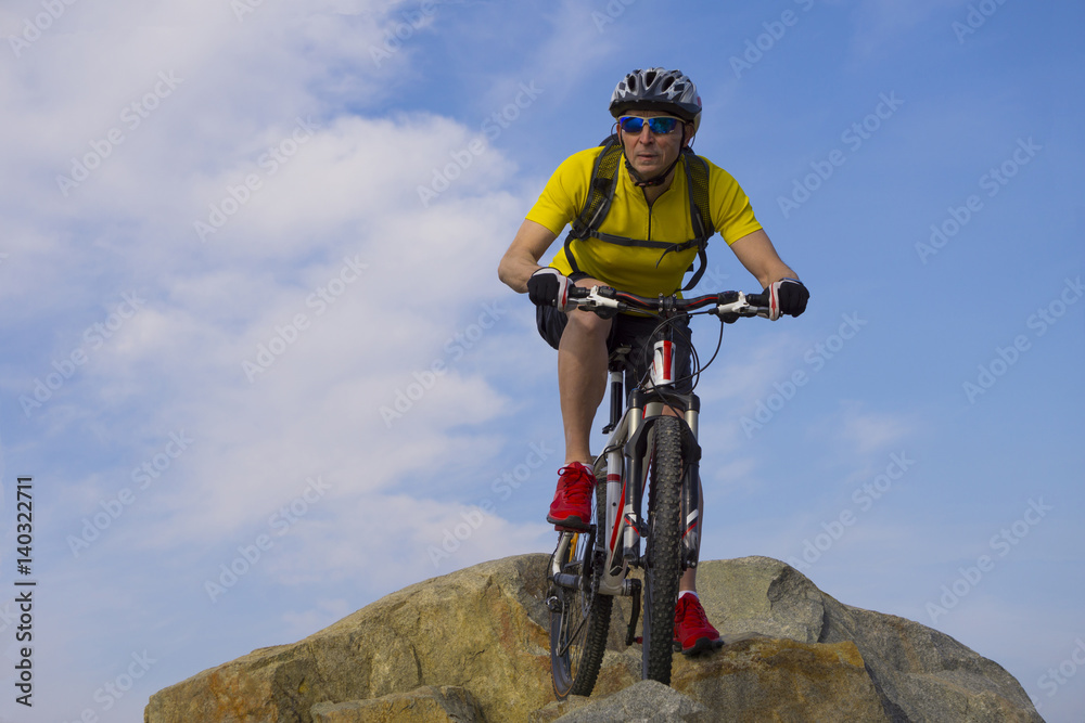 Fototapeta premium Downhill rider, in yellow t-shirt and red sneakers, on mountain bike with cloudy sky background